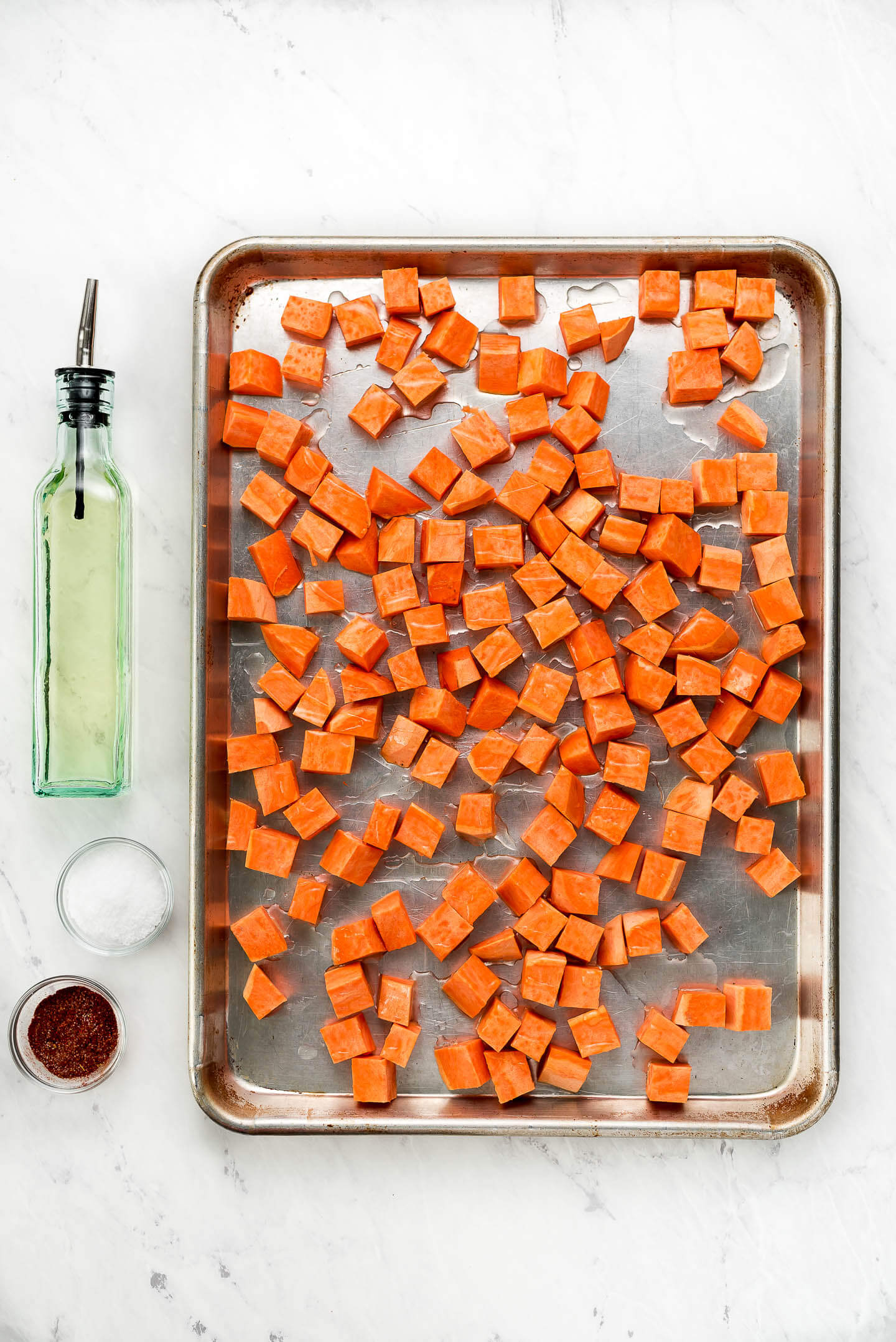 Cubed raw sweet potatoes on a baking sheet drizzled with olive oil and seasonings in small bowls to the side.