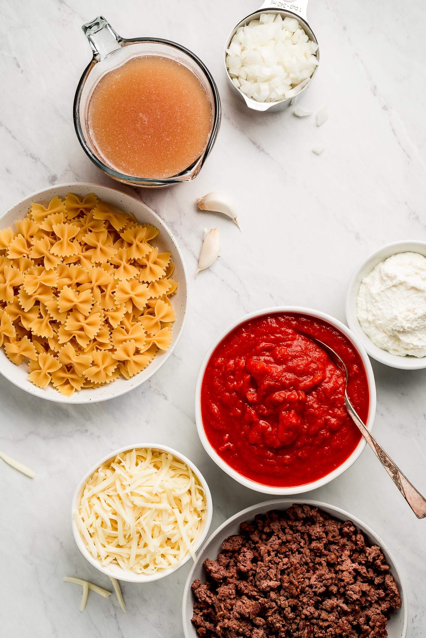 Ingredients on a marble surface in bowls-noodles, marinara sauce, ground beef, shredded cheese, broth, ricotta, onions.