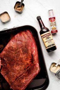 Ingredients on a marble surface- brisket, spices, Worcestershire sauce, and liquid smoke.